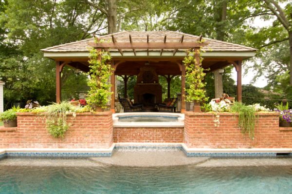 A serene vaulted cabana featuring a rustic stone fireplace and picturesque arbor, nestled by a sparkling swimming pool.