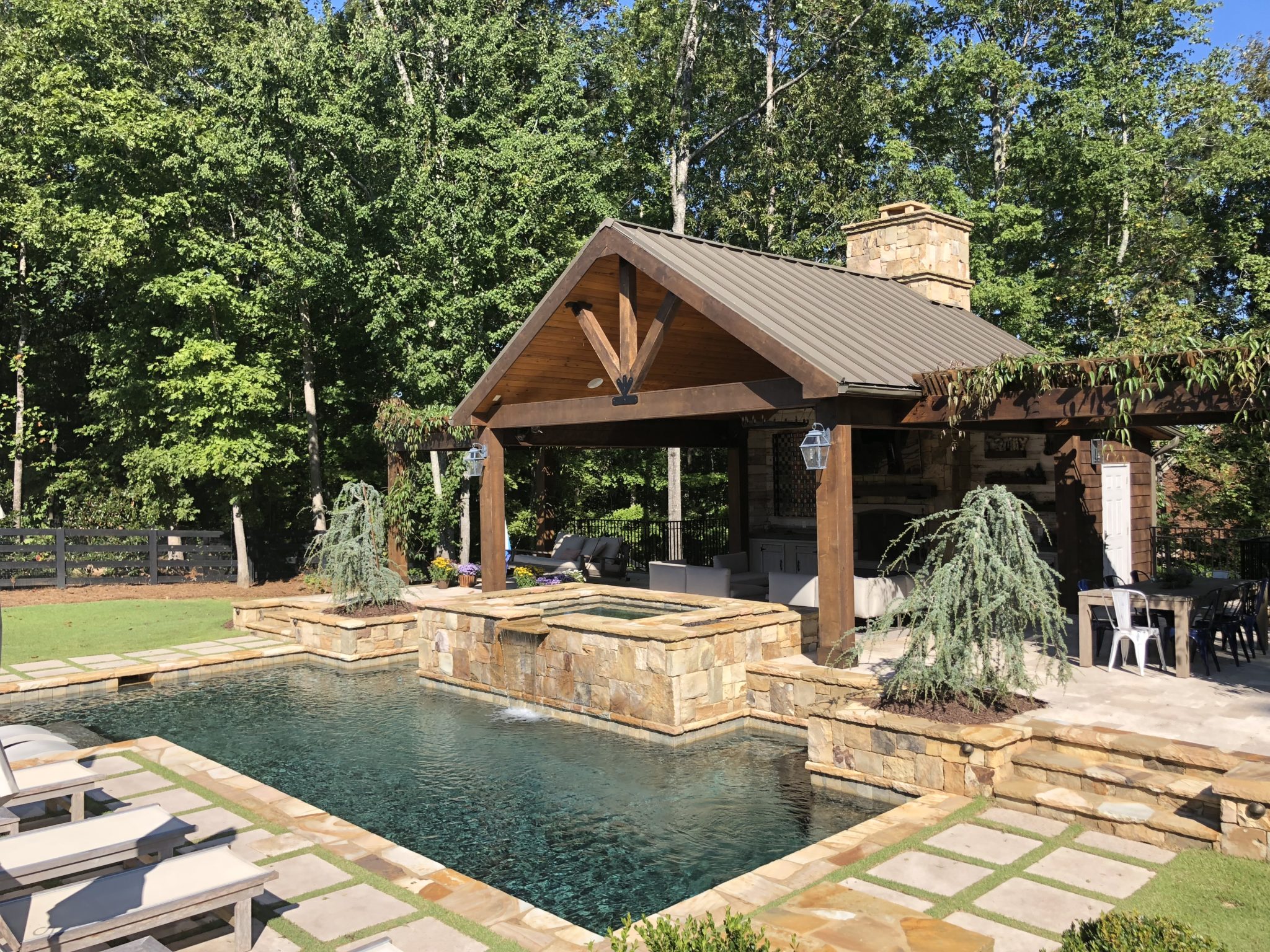 A serene vaulted cabana featuring a rustic stone fireplace and picturesque arbor, nestled by a sparkling swimming pool.