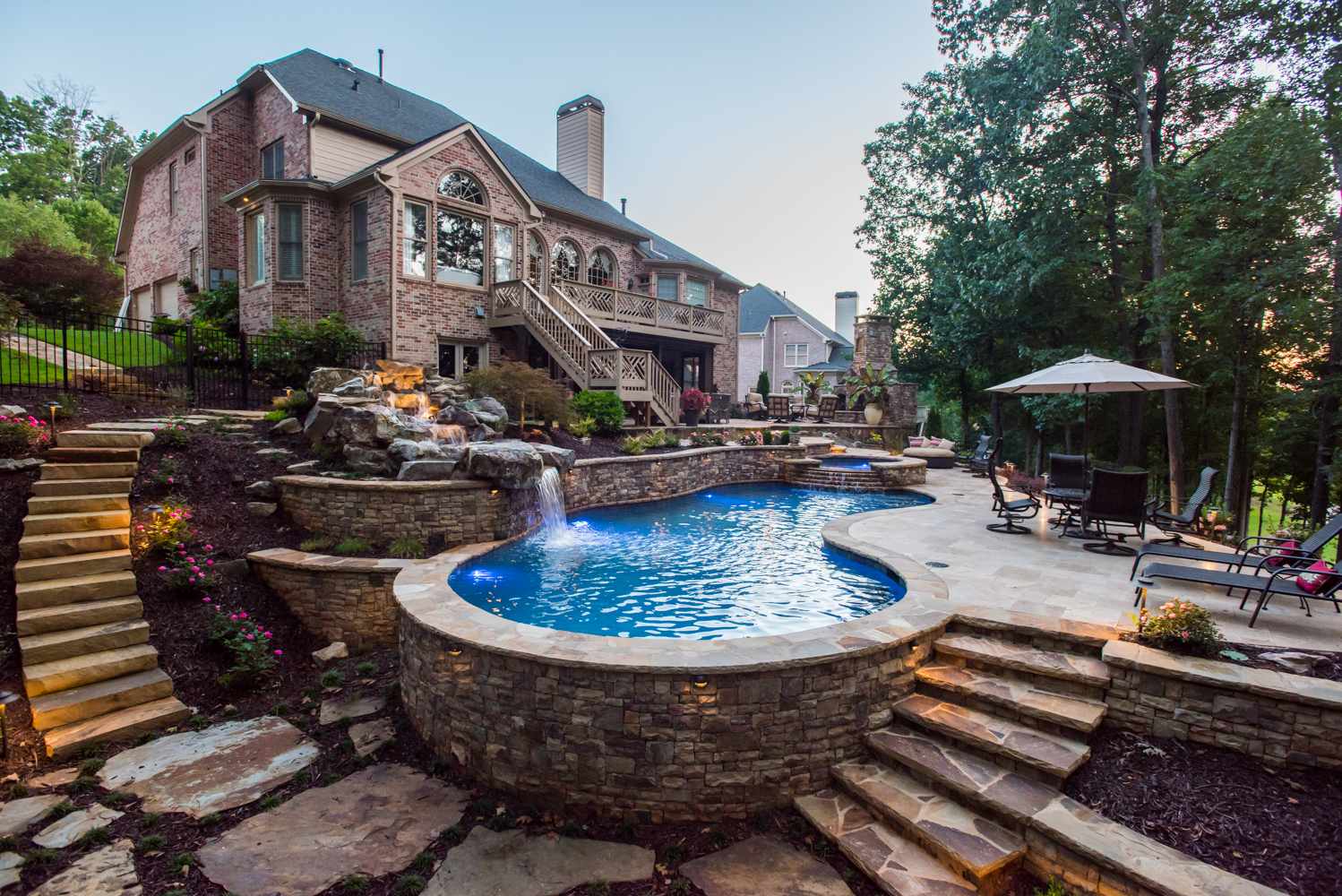 A custom freeform pool surrounded by lush greenery, with a boulder waterfall and flagstone steppers guiding to a seating area.
