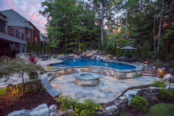 A breathtaking custom freeform pool surrounded by lush greenery, featuring a cascading boulder waterfall and a sunken fireplace for serene relaxation.