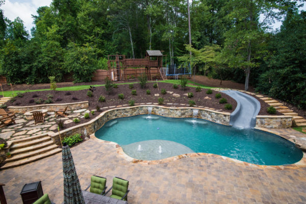 A custom freeform pool with a slide surrounded by lush greenery and lounge chairs.