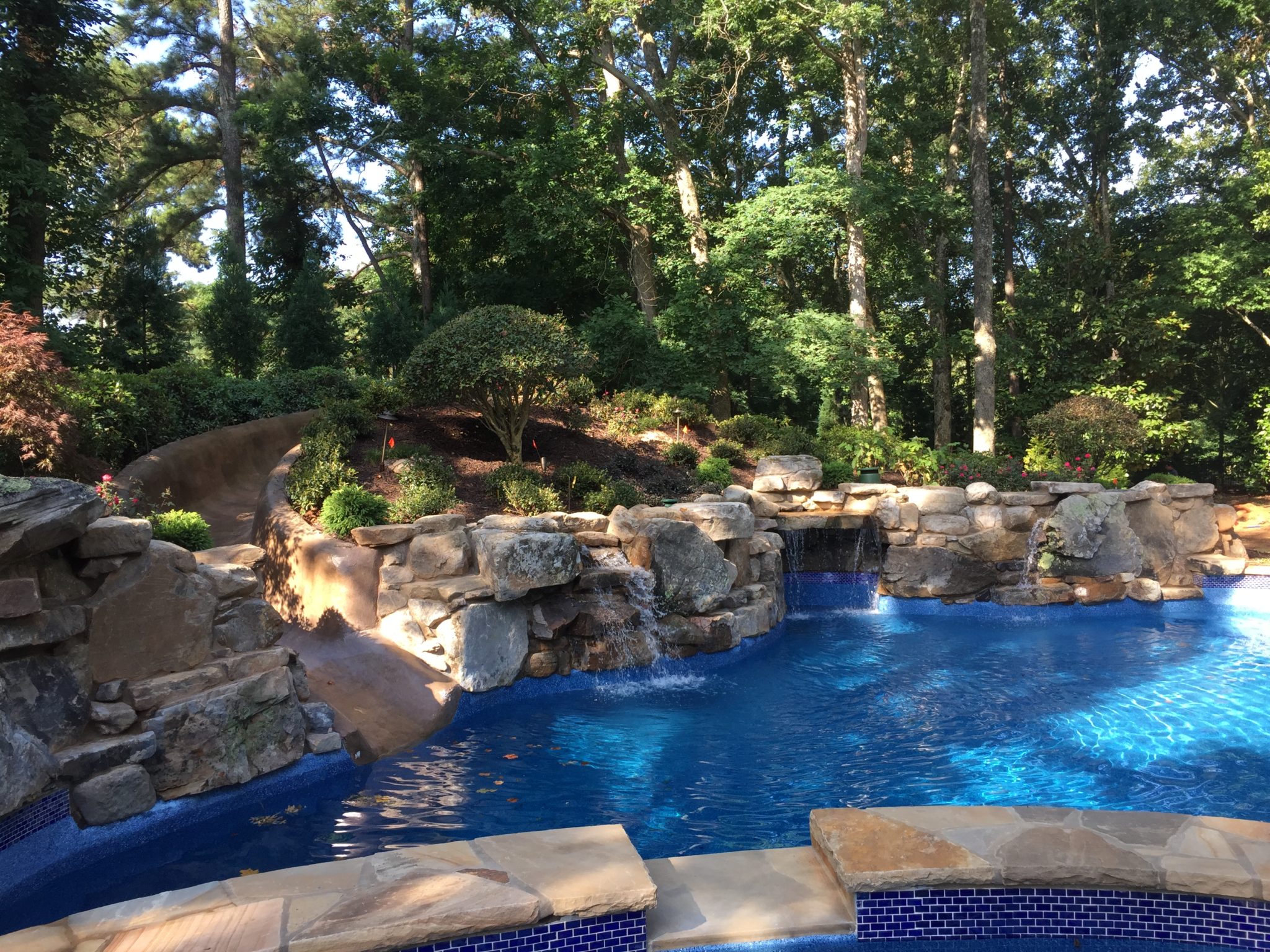 A custom freeform pool featuring a boulder waterfall grotto and slide surrounded by lush greenery and lounge chairs.
