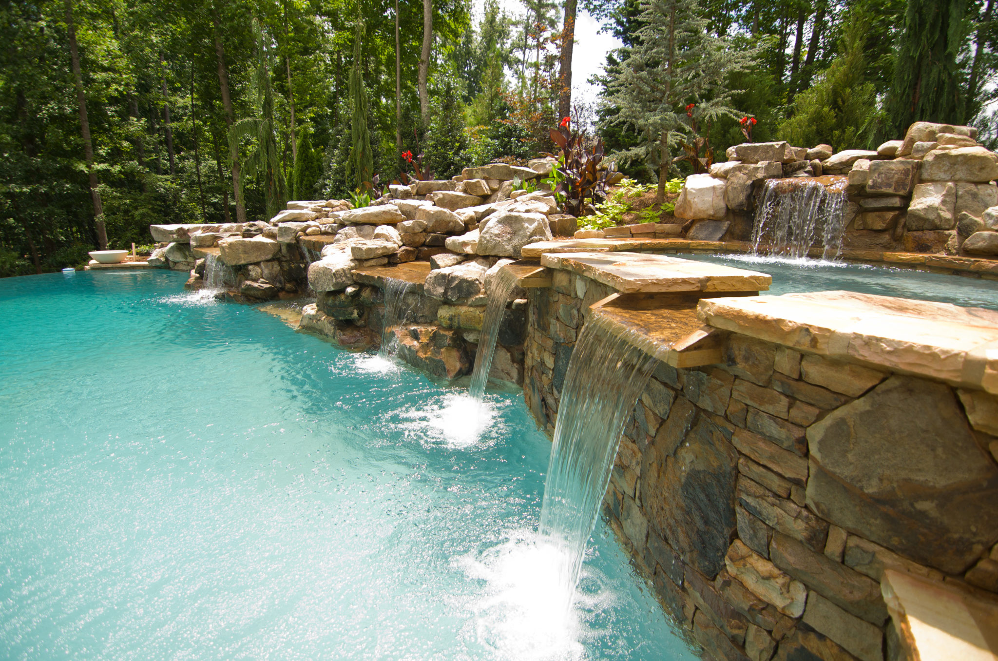 A custom freeform pool with a boulder waterfall spilling into a spa, surrounded by lush greenery and lounge chairs.