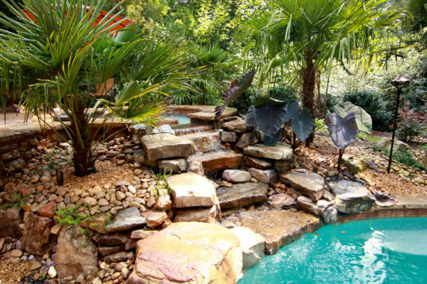 A serene spa cascades into a stunning three-tier boulder waterfall, surrounded by lush greenery.
