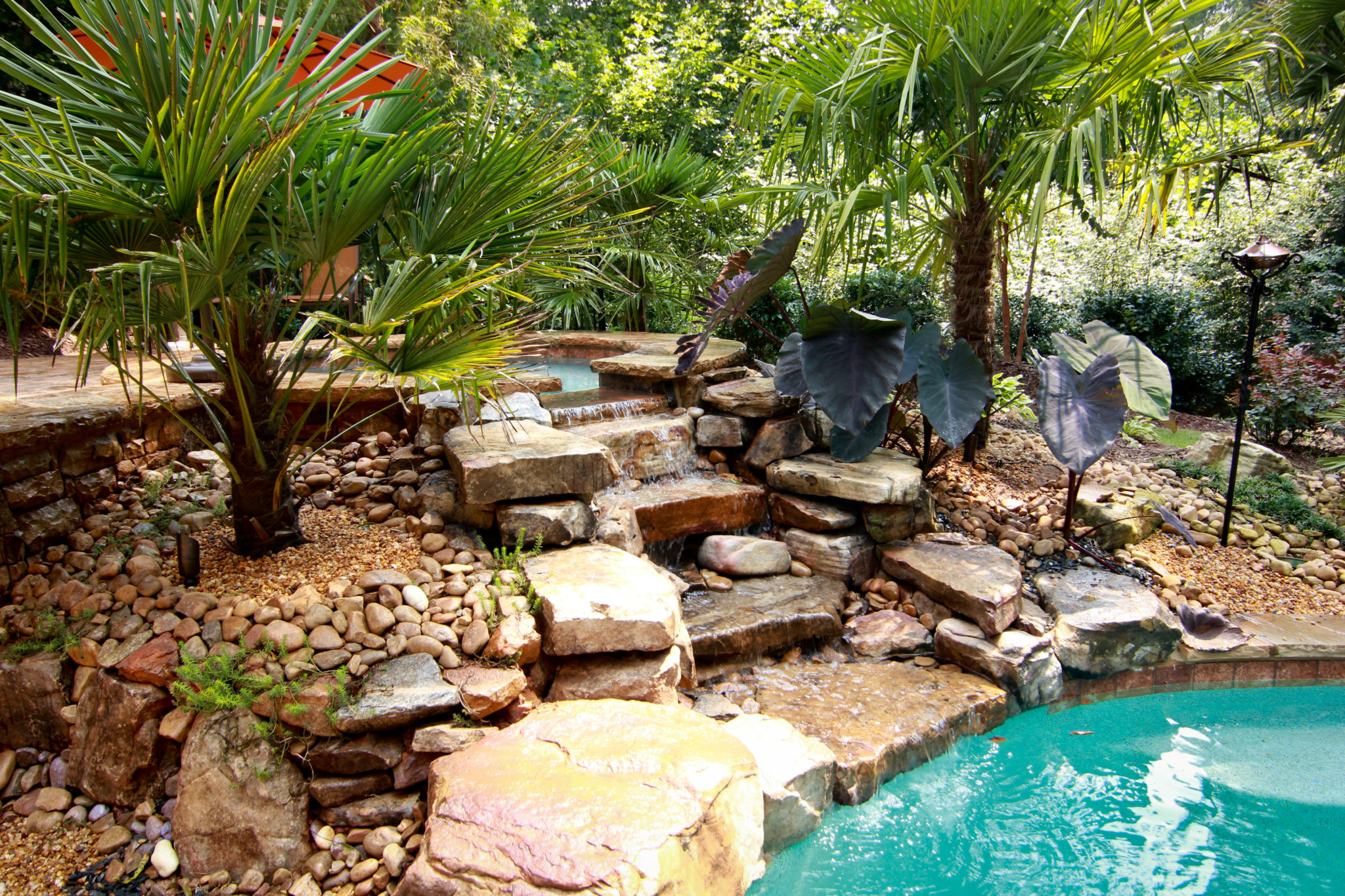 A serene spa cascades into a stunning three-tier boulder waterfall, surrounded by lush greenery.