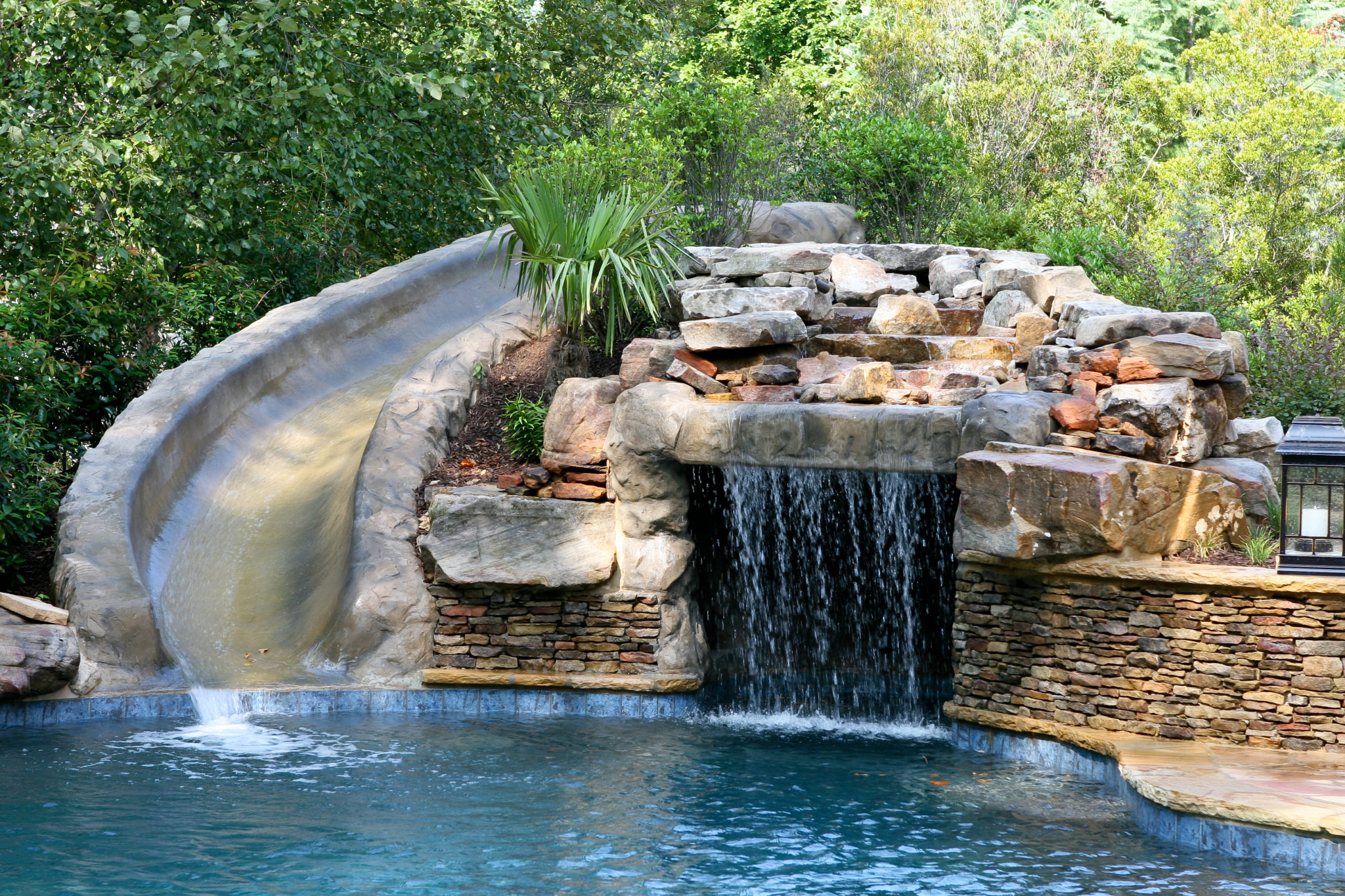 A custom boulder waterfall with a slide and grotto, surrounded by lush greenery.