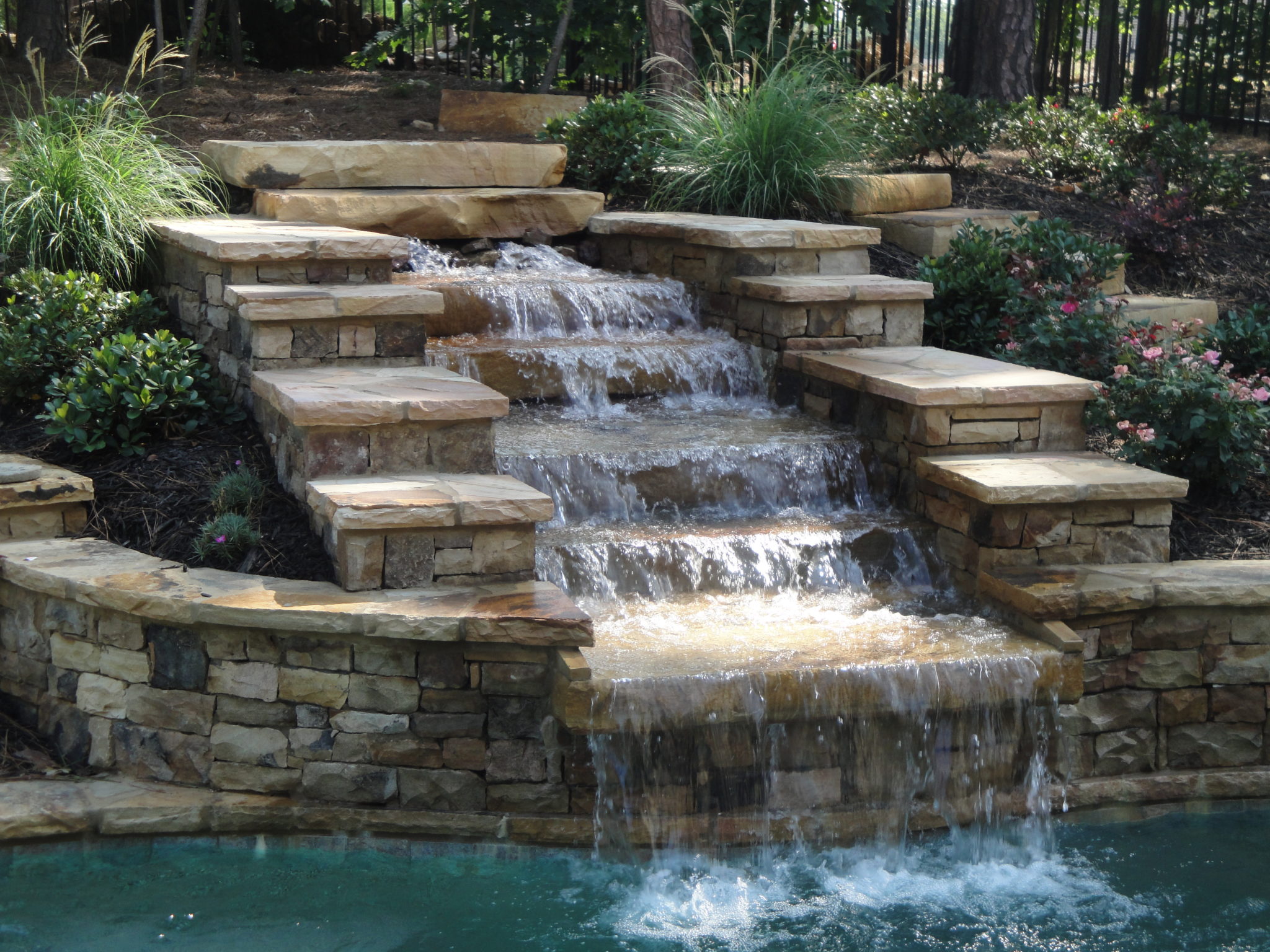 A five-tier stone waterfall cascading into a swimming pool.