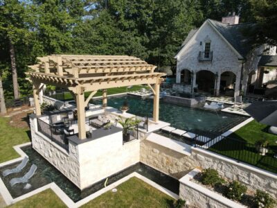Expansive vanishing edge pool with a 360-degree spa, accompanied by a vast pergola, outdoor kitchen, lush artificial turf, and meticulously designed custom landscape