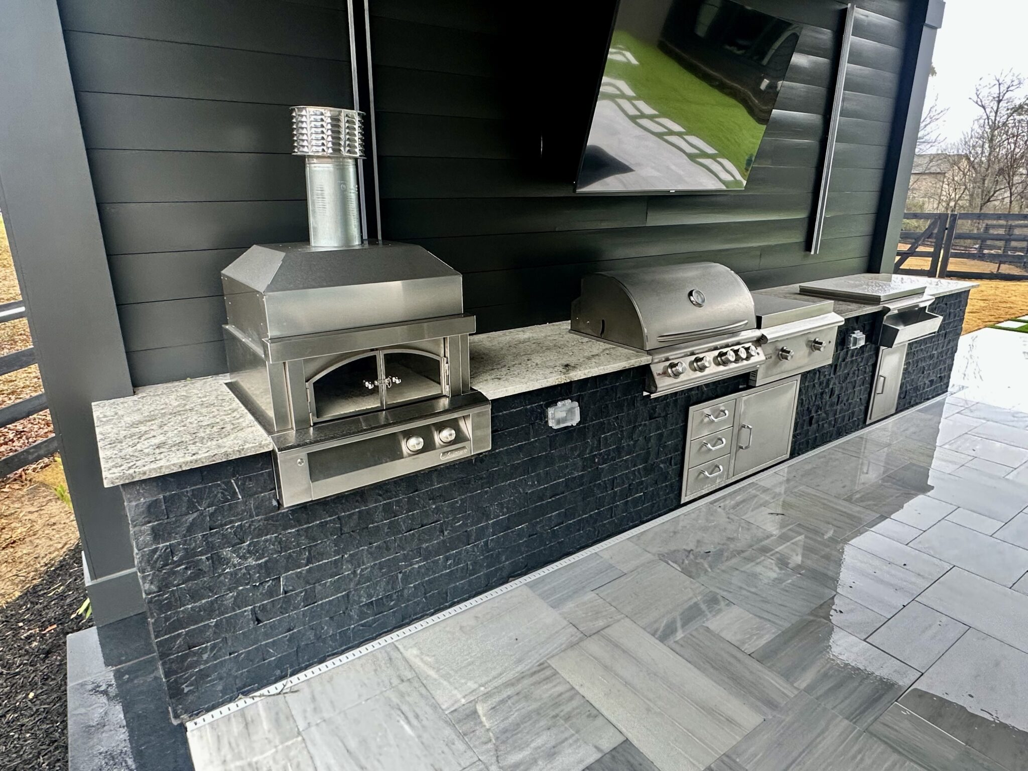 Modern outdoor kitchen with a pizza oven, grill, fridge, sink, and television under a cabana
