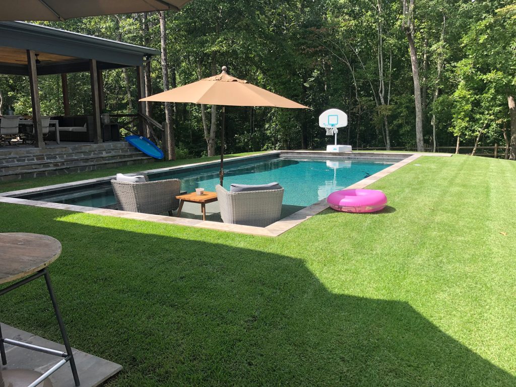Classic rectangular pool with tanning ledge loungers, patio umbrella, and stone coping in a wooded backyard